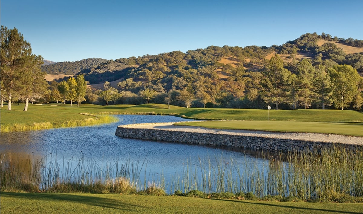 A manicured golf course is visible, featuring a green expanse alongside a tranquil waterway. The scene is framed by lush trees and rolling hills, creating a serene landscape. The soft silhouette of the hills enhances the peaceful atmosphere of the setting.