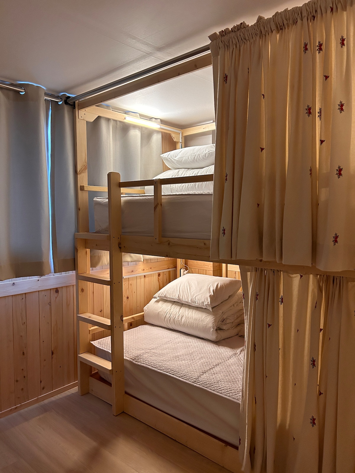 A shared room featuring two bunk beds with soft bedding, surrounded by light-colored wooden paneling. Curtains provide privacy for each sleeping area, while natural light filters through the large windows adorned with simple drapes.