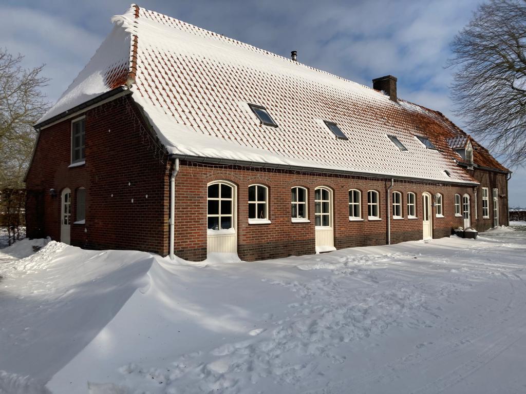 The snow-covered exterior of a spacious brick farmhouse is displayed, featuring a sloped roof with dormer windows. Multiple arched windows line the façade, reflecting a cozy atmosphere, while the surrounding snow creates a serene and peaceful environment.