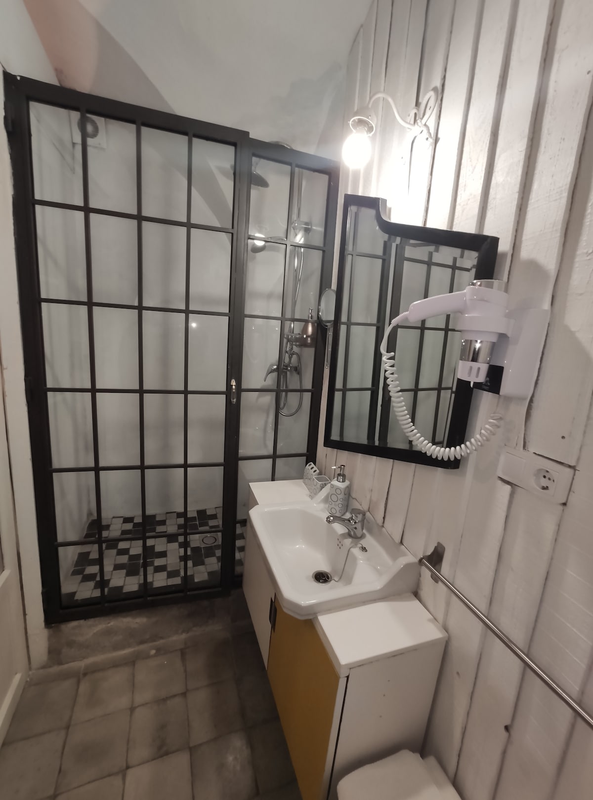 A modern bathroom features a glass-enclosed shower beside a sink with a mirror above. The floor displays patterned tiles, while light is provided by a wall fixture. The walls are adorned with wood paneling, contributing to a clean and functional design.