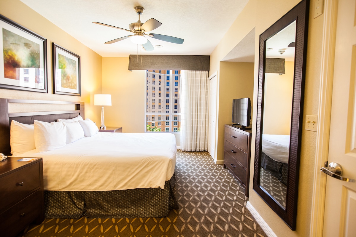 A spacious bedroom is furnished with a king bed, accented by soft white linens. Natural light fills the room through a large window, complemented by a mirror that reflects the inviting decor. A ceiling fan adds a touch of comfort to this relaxing space.