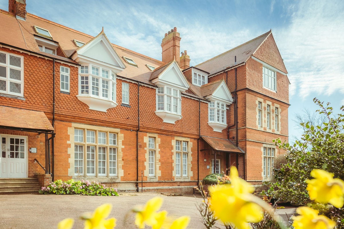 A striking red-brick building with intricate detailing is captured against a bright sky, featuring multiple gabled roofs and large windows. Colorful flowers in the foreground add vibrancy to the scene, highlighting the inviting exterior of the former school for gentlemen.