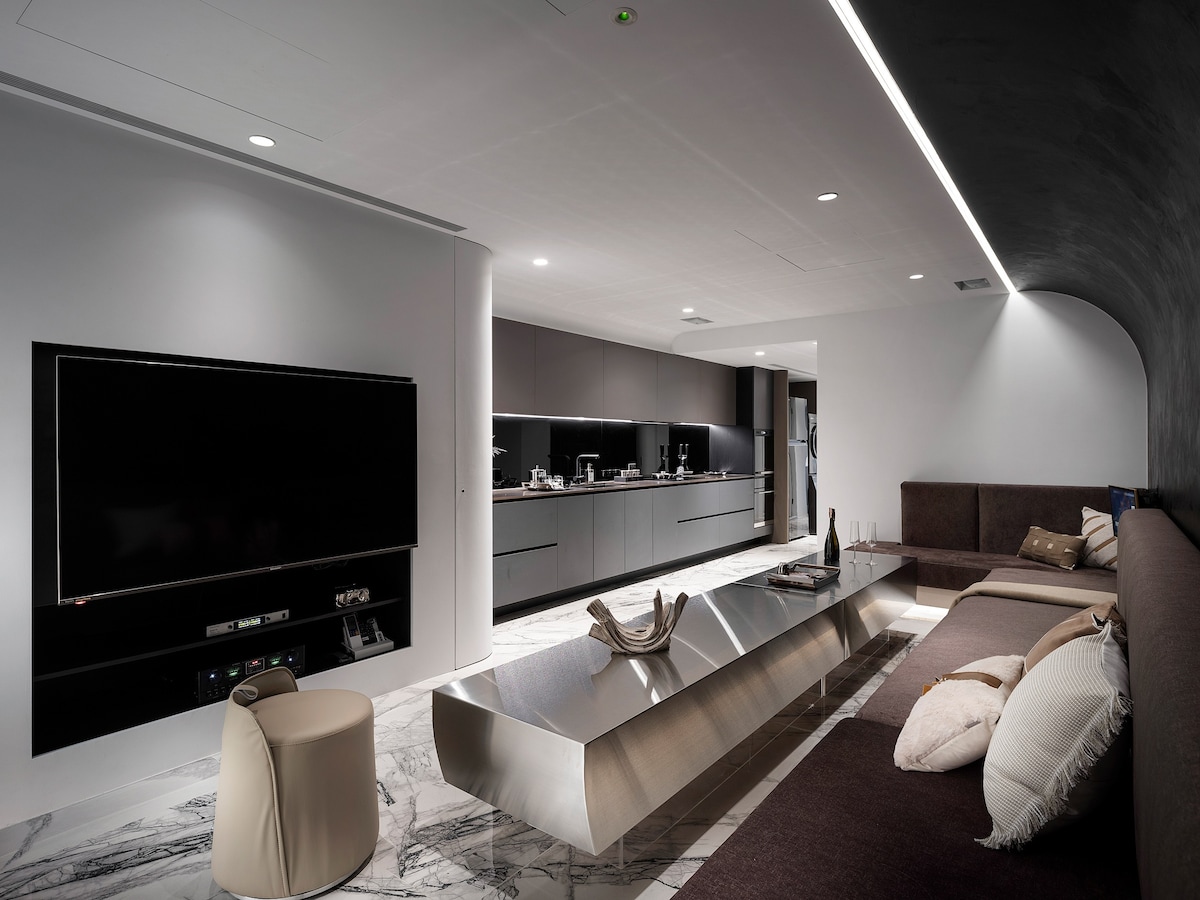 A modern living area features a sleek metallic coffee table centered on marble flooring. A long, low sofa spans one side, complemented by plush cushions. An expansive kitchenette, equipped with contemporary appliances, is visible in the background, creating a stylish and functional space.
