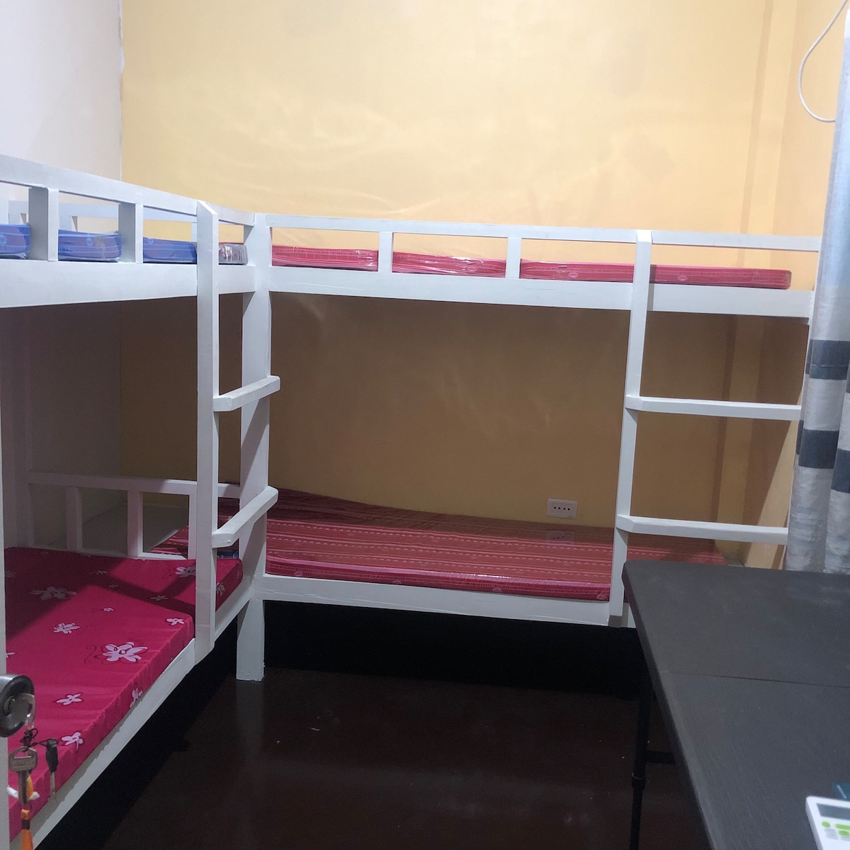A compact bunk bed arrangement is displayed, featuring two levels with bright red bedding and a simple white frame. A small table is positioned nearby, and the walls are painted a warm yellow, contributing to a cheerful ambiance.