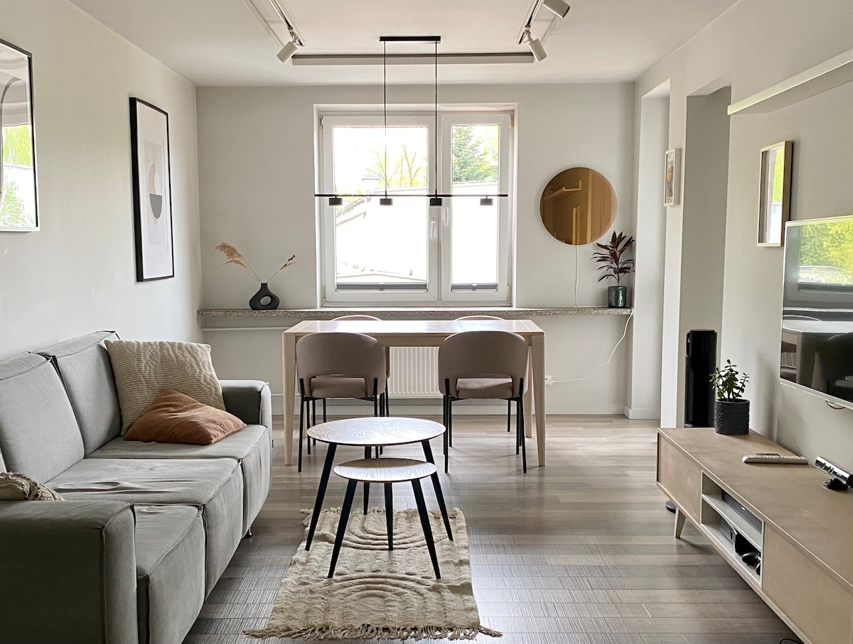 A modern living area is presented, featuring a light grey sofa with a decorative pillow. A wooden dining table with two chairs is positioned adjacent to a large window, allowing natural light to fill the space. A round coffee table rests on a textured rug, with a wall-mounted television nearby.