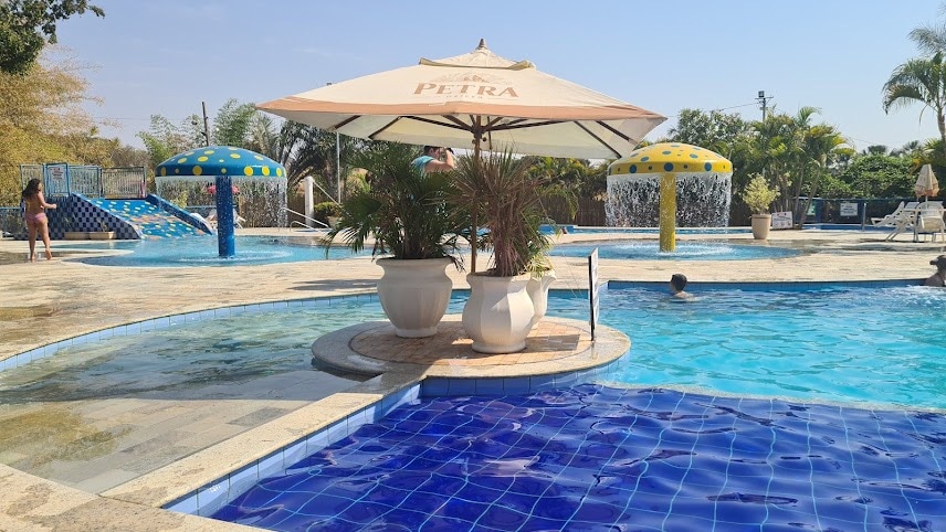 A serene pool area is framed by vibrant landscaping and features multiple water slides. The clear water reflects sunlight, creating a welcoming ambiance. An umbrella provides shaded seating next to a large plant pot, enhancing the outdoor leisure space.