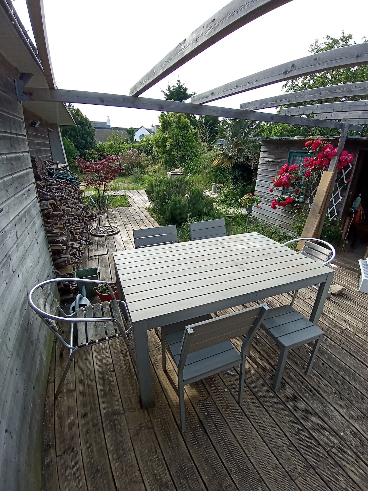 A wooden terrace is surrounded by a lush garden, featuring a dining table with matching chairs. Vibrant flowers and shrubs add a natural touch, while a trellis supports climbing plants. The setting offers a calm outdoor space, ideal for family gatherings or relaxation.
