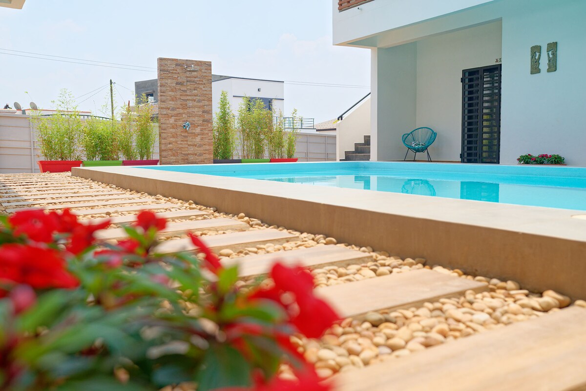 An outdoor area features a clear swimming pool bordered by smooth stones and vibrant flower beds. Decorative plants provide a green backdrop, while clean lines and modern architecture contribute to a minimalist aesthetic.
