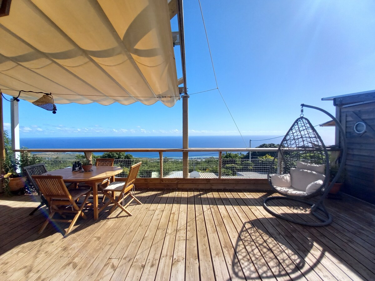A spacious outdoor deck features a dining table surrounded by chairs and a hanging swing chair. The panoramic view of the ocean is visible in the background, with clear blue skies and a gentle awning providing shade.