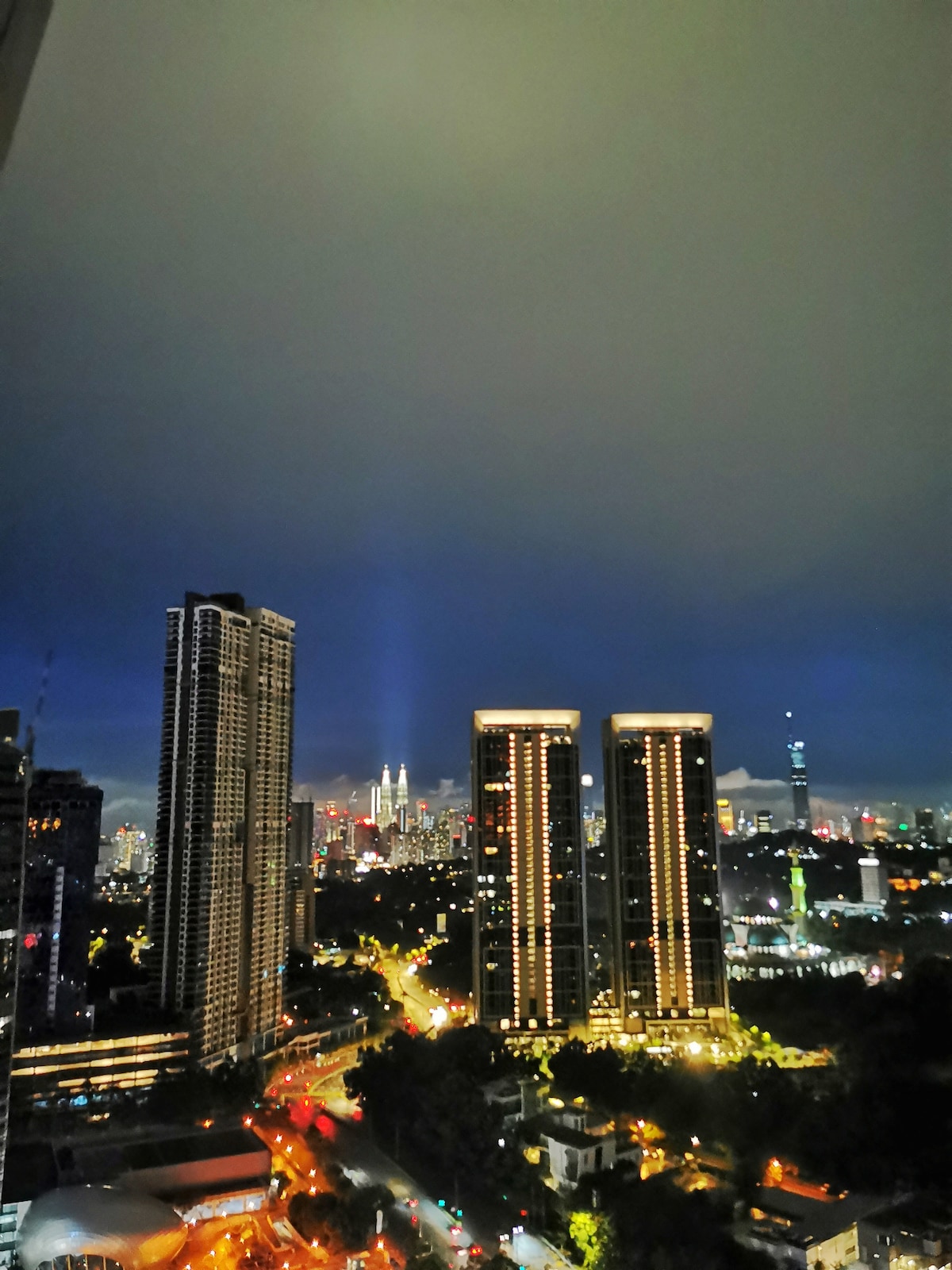 A nighttime cityscape showcases towering buildings bathed in ambient lights. The skyline features prominent structures, including the Petronas Towers, which stand out against a cloudy sky. Streets below are illuminated, revealing the movement of vehicles and the vibrancy of the urban environment.