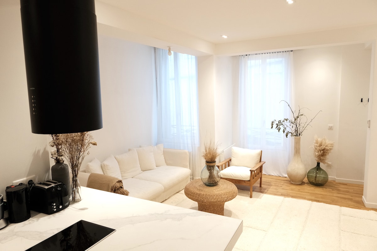 A bright living area features a white sofa and a light-toned armchair, complemented by a round coffee table. Natural light filters through sheer curtains, illuminating decorative plant arrangements in vases on the floor. A soft, textured rug covers the wooden flooring.