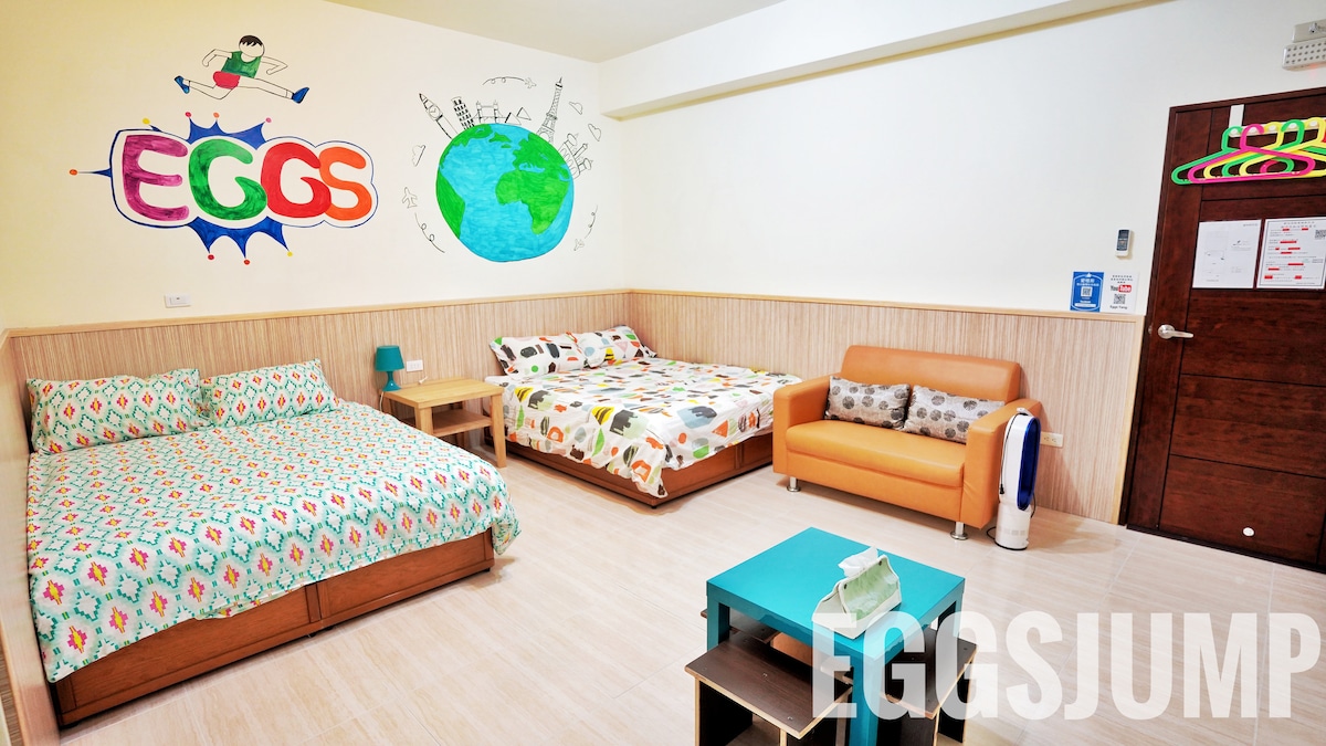 A colorful guest room features two spacious beds with unique bedding patterns. A small wooden table sits in the center with an orange sofa nearby. Whimsical wall art depicting playful characters and a globe adds a lively touch to the atmosphere.