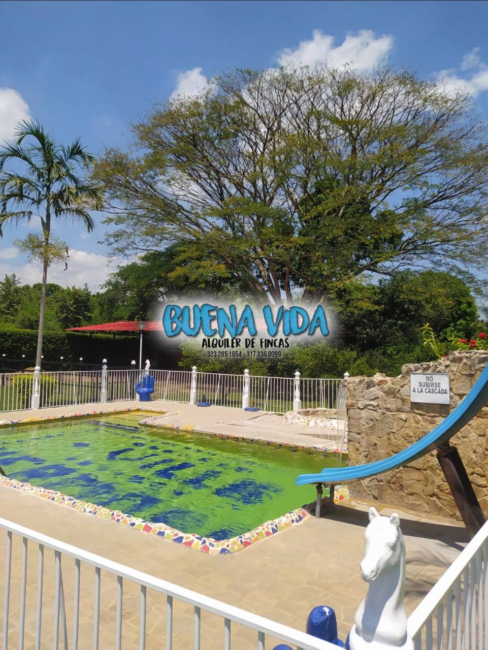 A large outdoor swimming pool is highlighted, featuring a colorful tiled border and a water slide on one side. Lush greenery surrounds the pool area, under a clear blue sky, while a spacious terrace offers additional seating options nearby.