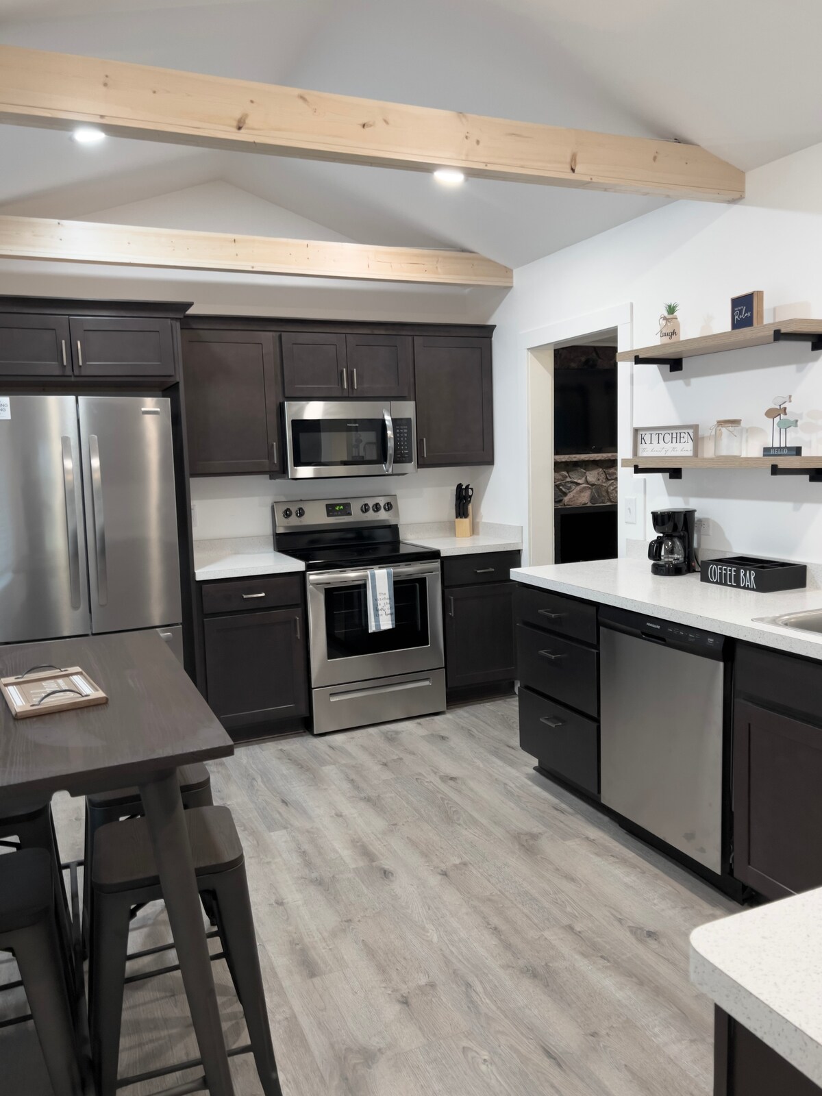 The kitchen features dark cabinetry paired with modern stainless steel appliances, including a refrigerator, oven, and dishwasher. A countertop with a coffee bar setup and seating for four complements the airy atmosphere created by the vaulted ceiling and warm wood beams.