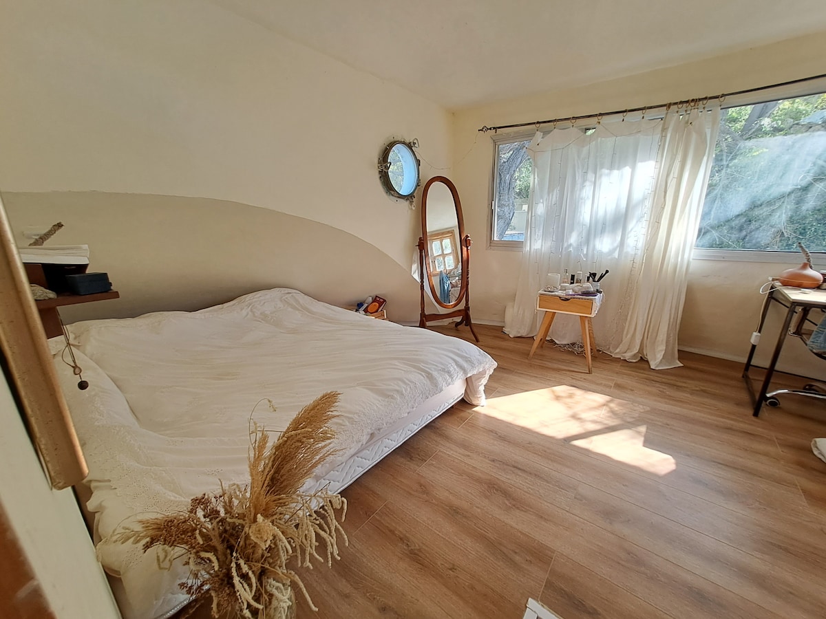 A bright sleeping area features a spacious bed covered with a white duvet. Natural light pours in through large windows, illuminating the warm wooden floor. A standing mirror and a small table are positioned nearby, adding functional elements to the room.