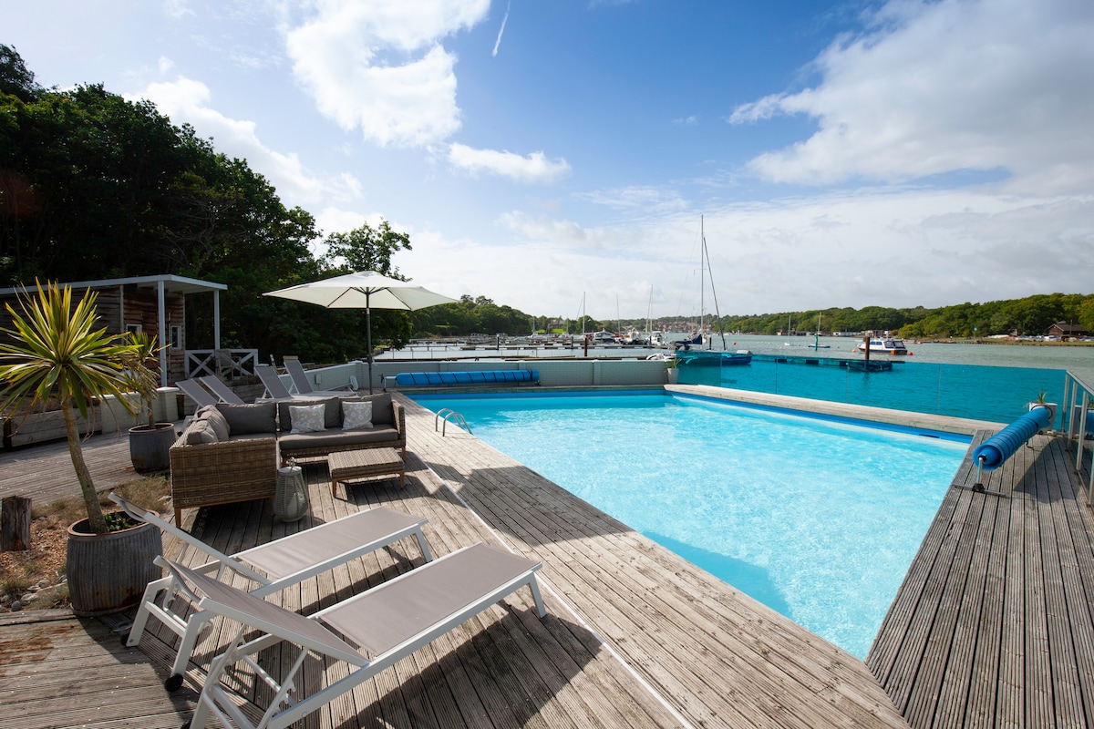 A large swimming pool surrounded by a spacious wooden deck is visible, with loungers and seating areas for relaxation. Umbrellas provide shade, and the vibrant water of the creek is seen in the background, along with boats moored nearby. Lush greenery frames the area.