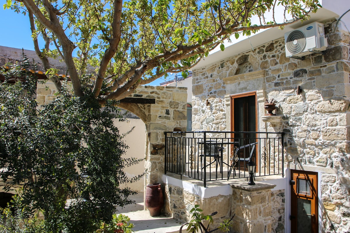 A traditional stone house features a balcony adorned with a black railing, offering outdoor seating. Lush greenery surrounds the space, with a large tree casting shade. An air conditioning unit is mounted on the exterior wall, contributing to comfort. Decorative pottery enhances the courtyard ambiance.