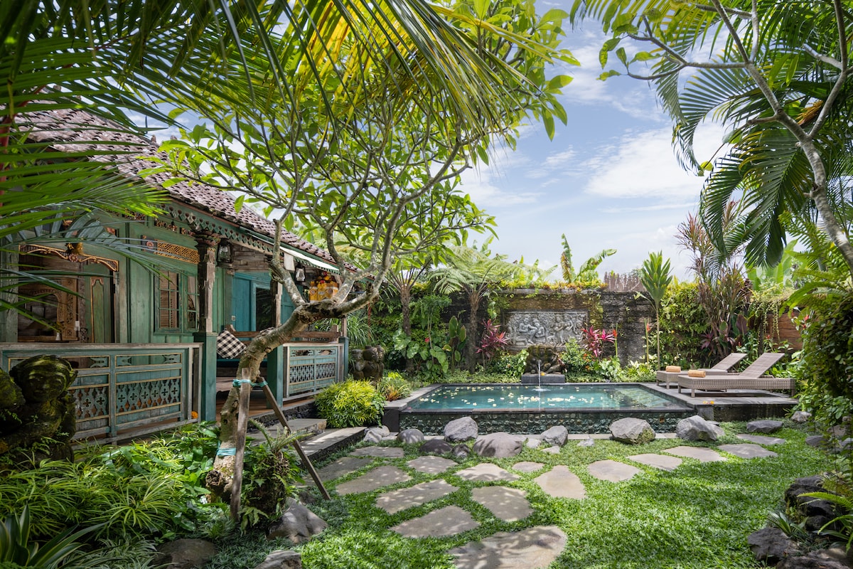 A serene garden setting is revealed, featuring a sparkling pool surrounded by lush greenery. A traditional Balinese-style structure is visible, framed by tropical plants and trees. Natural stone pathways lead through the vibrant landscape, emphasizing the tranquility of the outdoor space.
