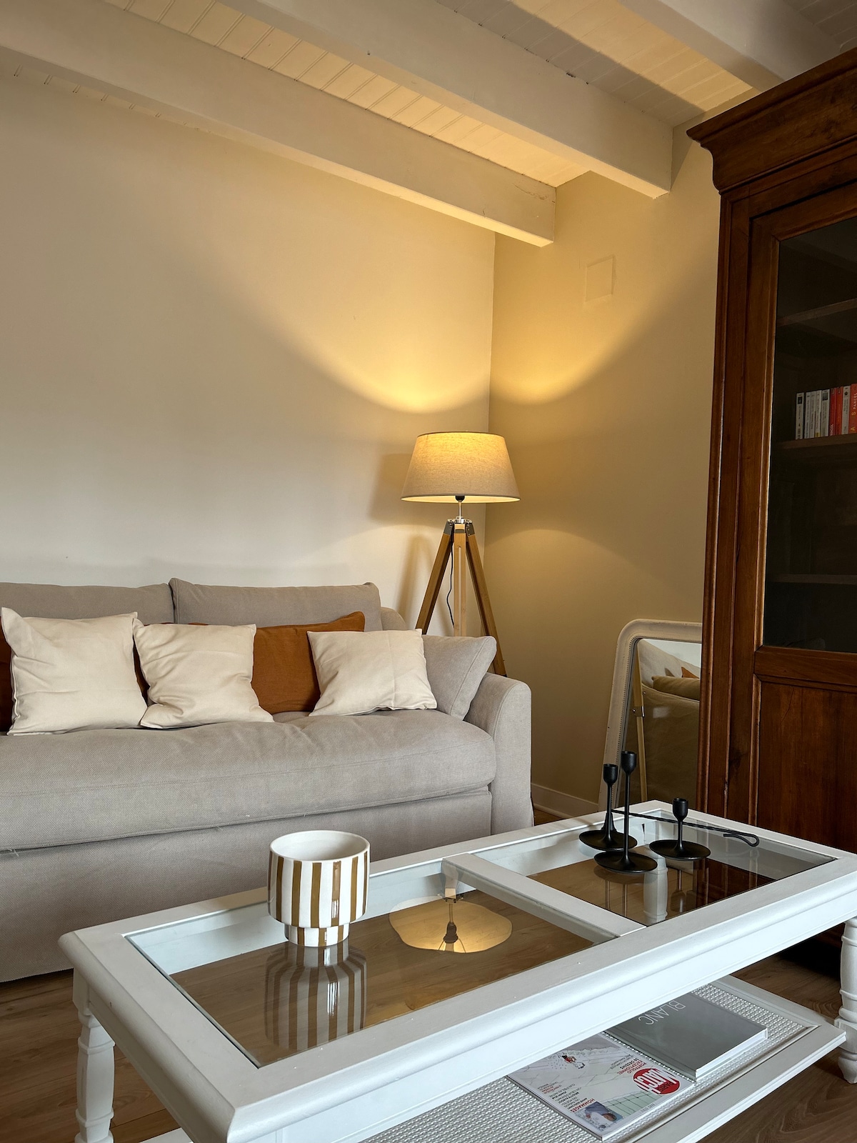 The living area features a light-colored sofa adorned with decorative pillows. A stylish floor lamp casts a warm glow, and a glass-top coffee table displays a decorative bowl and two candles. A wooden cabinet and a mirror can also be seen in the space.