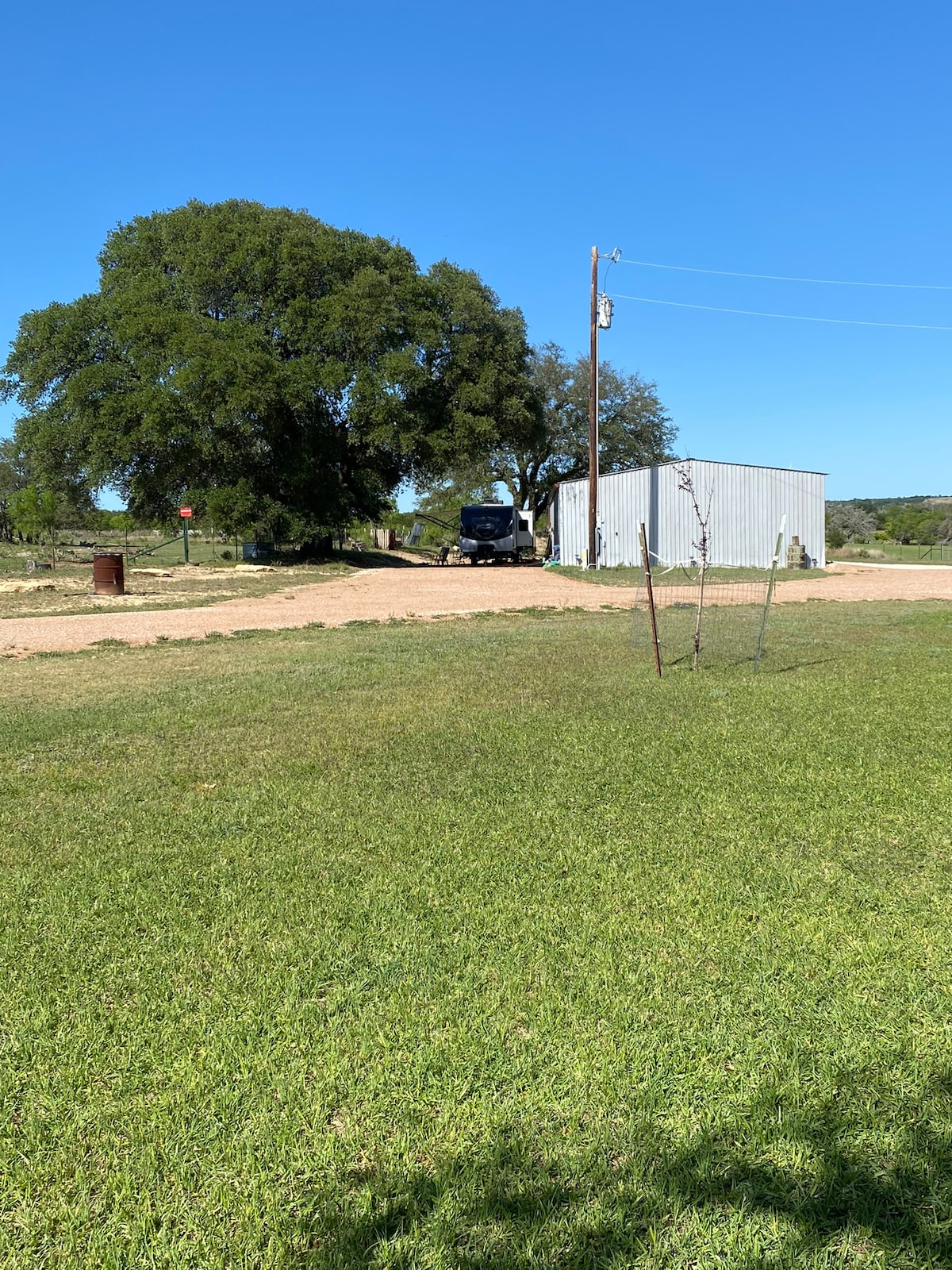 A grassy area extends towards a gravel road leading to a silver storage shed. A large oak tree provides shade nearby, while a parked RV is visible beyond the shed. The clear blue sky enhances the serene outdoor setting.