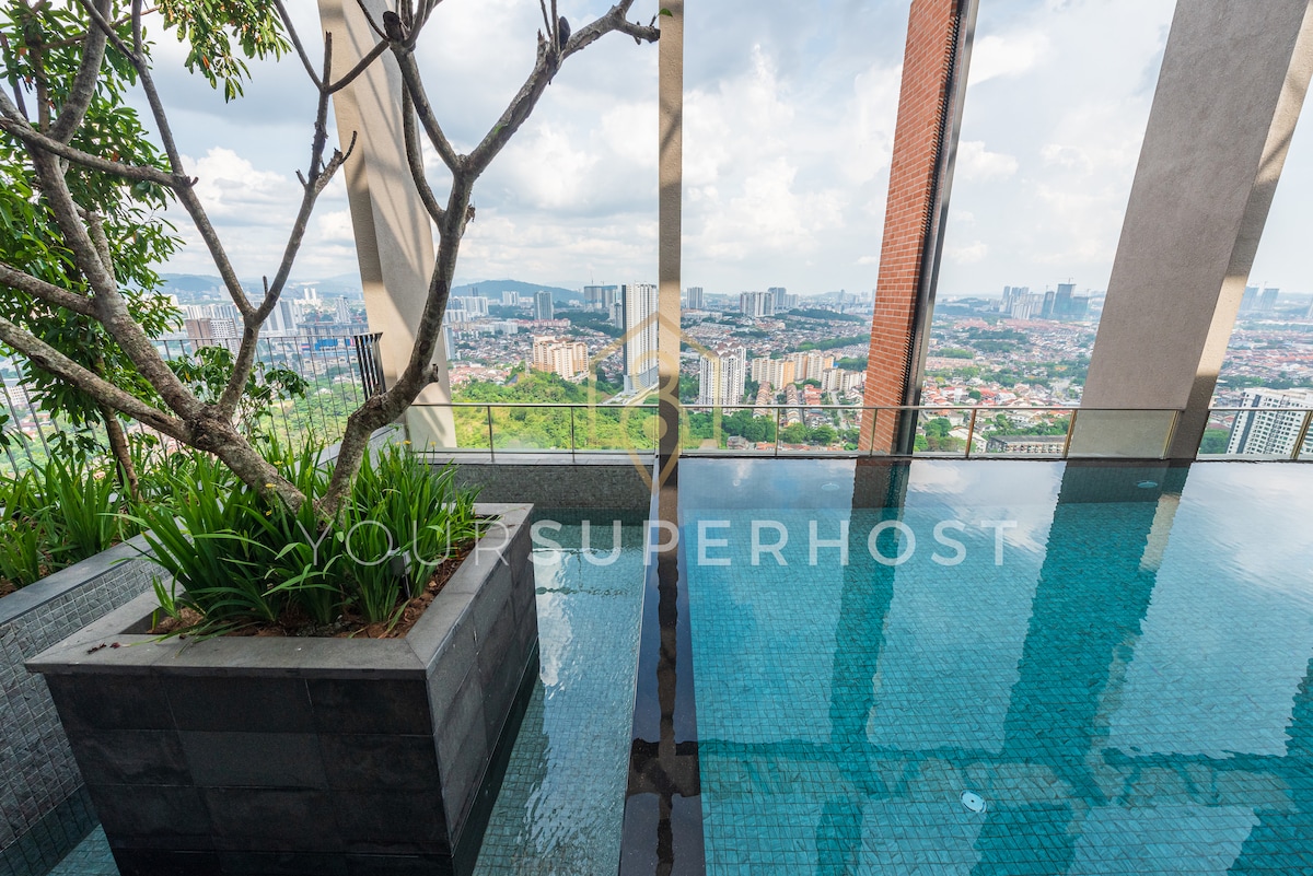 An infinity pool is situated on a deck with panoramic views of Kuala Lumpur's skyline. Lush greenery is visible along the edges, complemented by sleek stone features. The cityscape is showcased in the background beneath a partly cloudy sky.