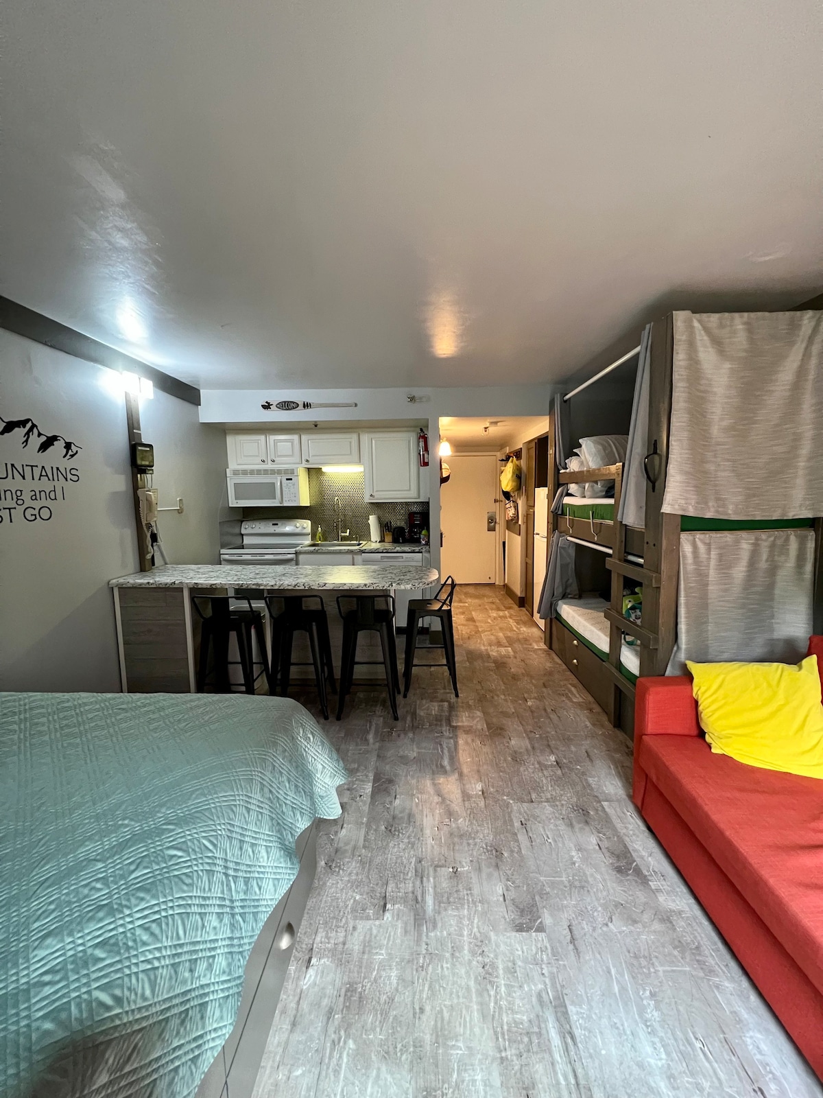 The studio features a combined living and kitchen area with a full kitchen and dining space. A spacious bed is positioned on the left, while a red couch and bunk beds are located on the right. Light-toned flooring enhances the open feel of the space.