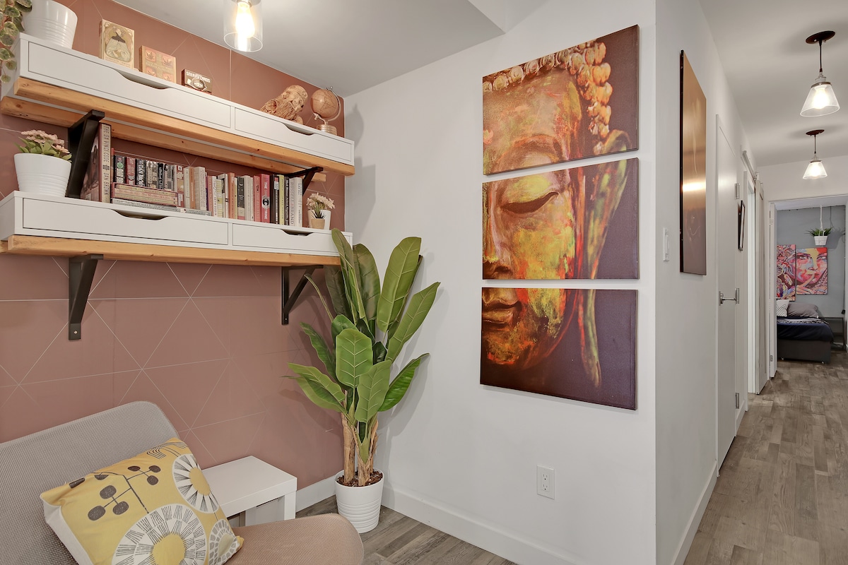 A cozy sitting area features a light-colored couch with a decorative pillow, complemented by a wooden shelf displaying books and decorative items. A potted plant adds a touch of greenery, while a wall art piece depicts a serene Buddha face, creating a calm ambience.