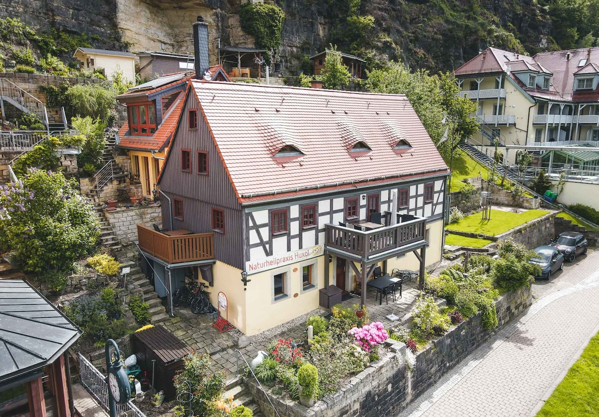 A charming half-timbered house is nestled against a rocky hillside, surrounded by lush greenery. A spacious terrace with outdoor seating is visible, along with flower beds featuring colorful blooms. The building's unique architecture adds character to the inviting exterior.