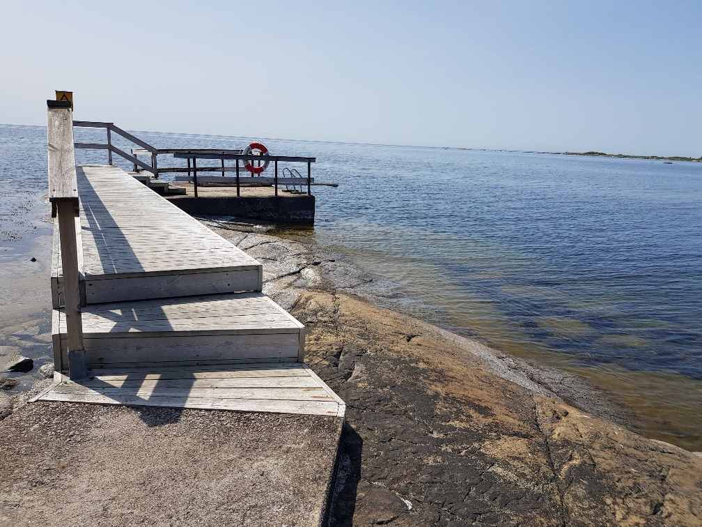 A wooden dock extends from the rocky shoreline into calm waters, providing access to the sea. Railings are present along the walkway, and a lifebuoy is visible on the dock. Sunlight reflects off the water's surface, creating a serene atmosphere.