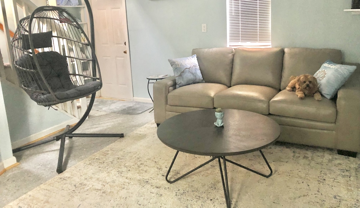 A cozy living area features a light-colored sofa adorned with accent pillows. A modern, circular coffee table sits centrally, complemented by a decorative item. A unique hanging chair is positioned nearby, with a small dog comfortably resting on the sofa.