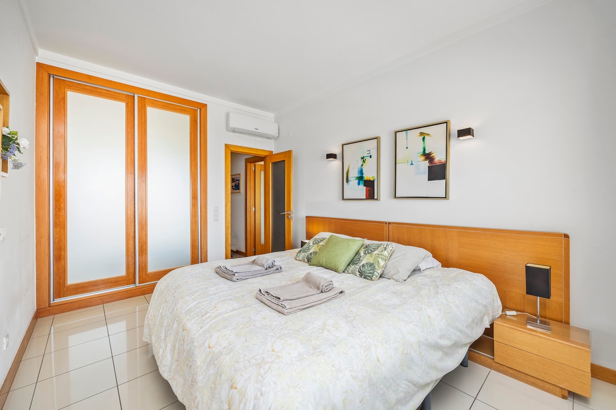 A spacious bedroom features a large bed with light bedding and neatly arranged towels. Two framed artworks adorn the wall above the bed, while wooden cabinetry offers ample storage. Natural light is provided through sliding glass doors leading to the hallway.