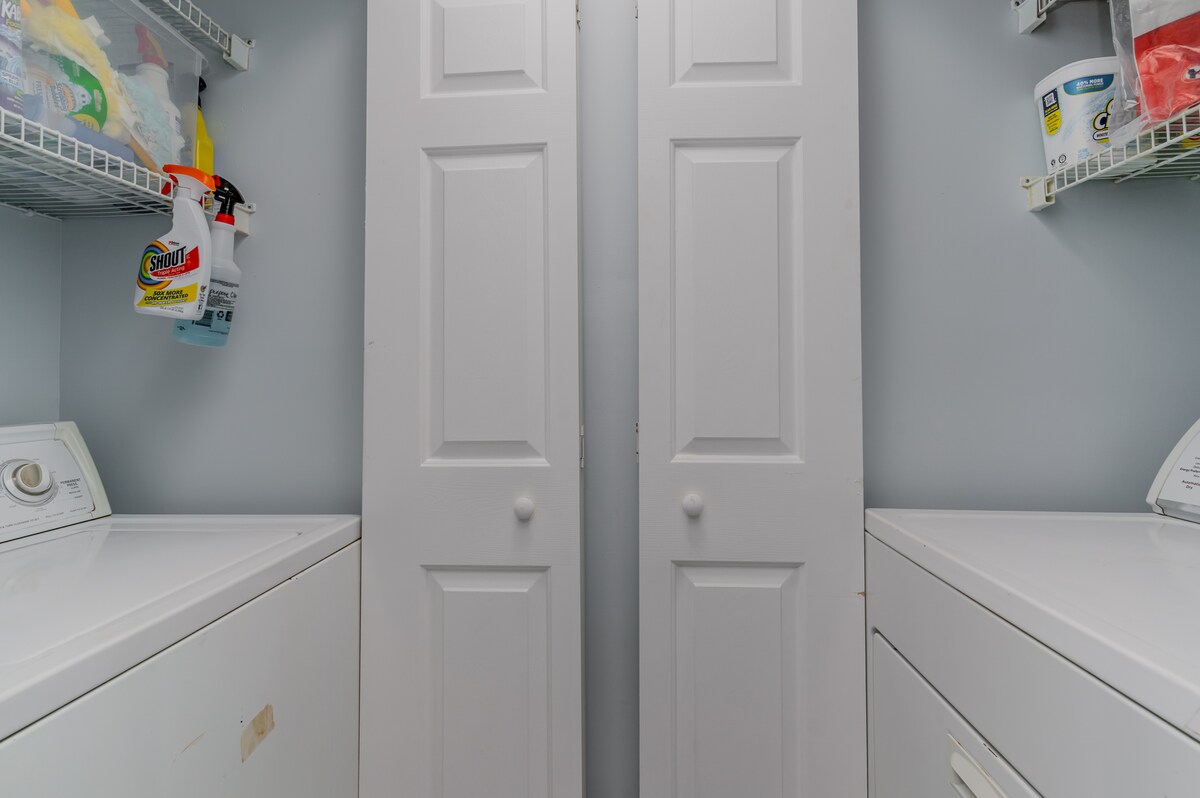 A laundry area features a stacked washer and dryer, positioned side by side. Bright shelving above provides storage for cleaning supplies and other essentials. The room is painted in a soft blue hue, enhancing the functional space.