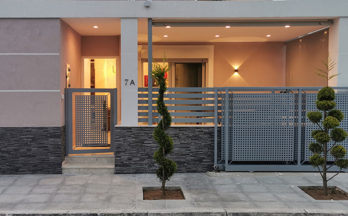 The entrance to the property is framed by modern landscaping, featuring neatly trimmed bushes and an ornamental fence. A well-lit pathway leads to the interior, with a welcoming ambiance created by subtle wall lighting that enhances the architectural design.