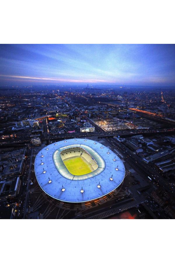 An aerial view captures the Stade de France stadium, illuminated under a twilight sky. Surrounding urban landscapes are visible, showcasing a mix of buildings and streets, while the vibrant green pitch stands out at the stadium's center, offering a glimpse of the bustling city beyond.