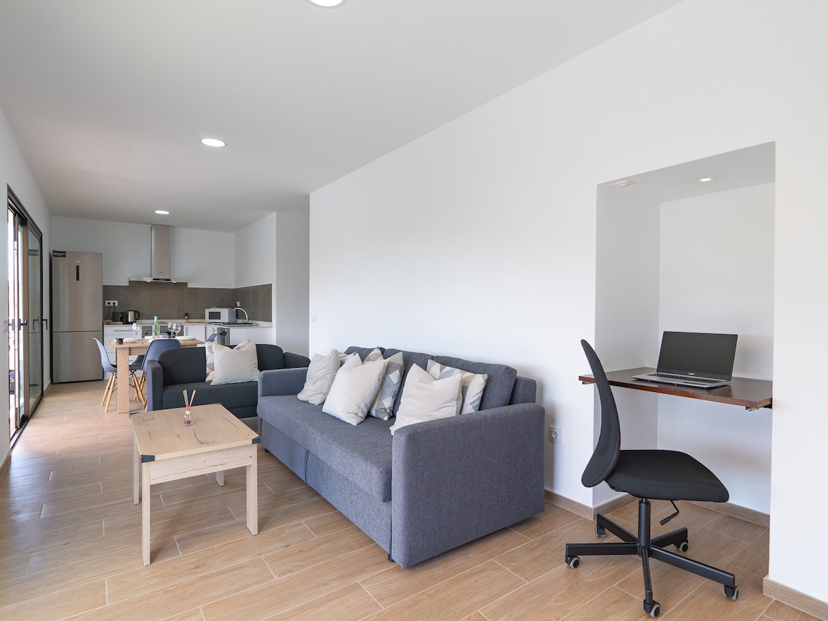 A spacious living area features comfortable seating with cushions and a wooden coffee table. A desk with a chair is positioned in a nook, providing a workspace. Natural light floods through large windows, enhancing the open layout that connects the living space to the kitchen.