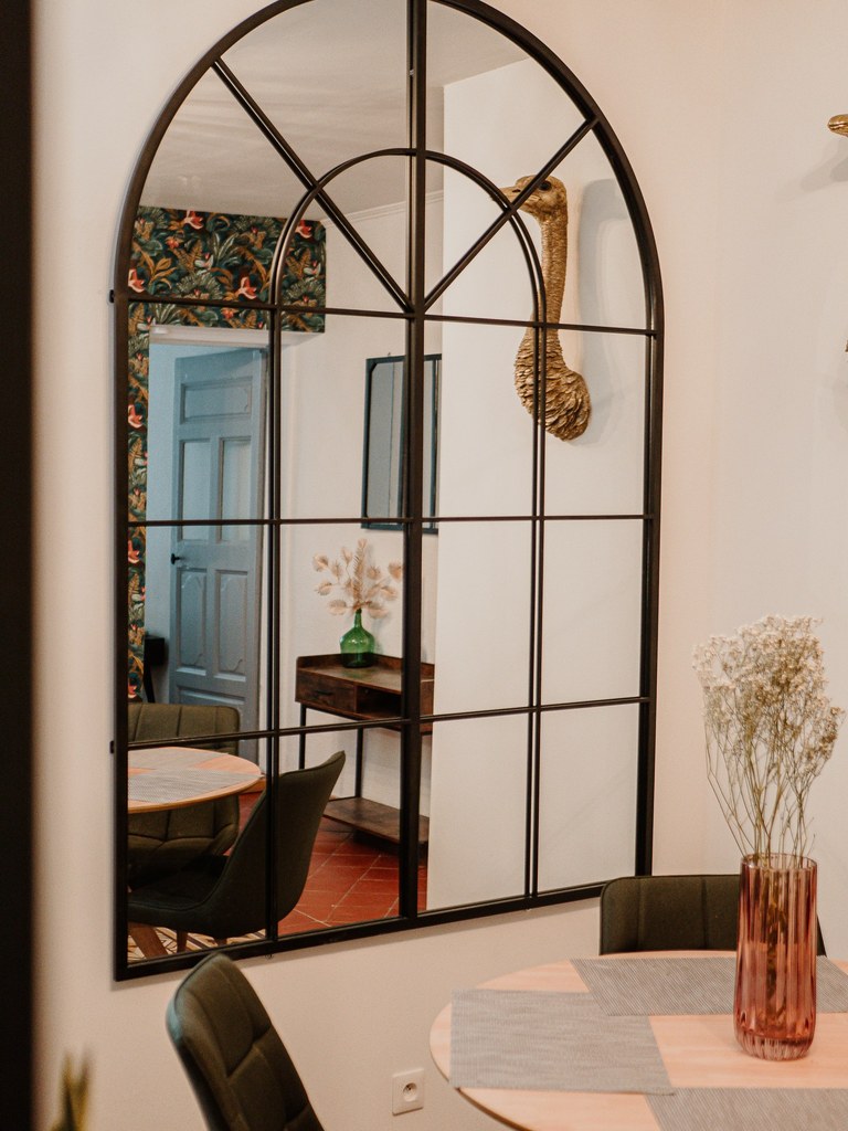 A stylish mirror with an arched design reflects the wall adorned with tropical foliage. A round dining table with four green chairs is visible in the foreground, accompanied by a floral arrangement in a tall vase. A wooden console table rests nearby.