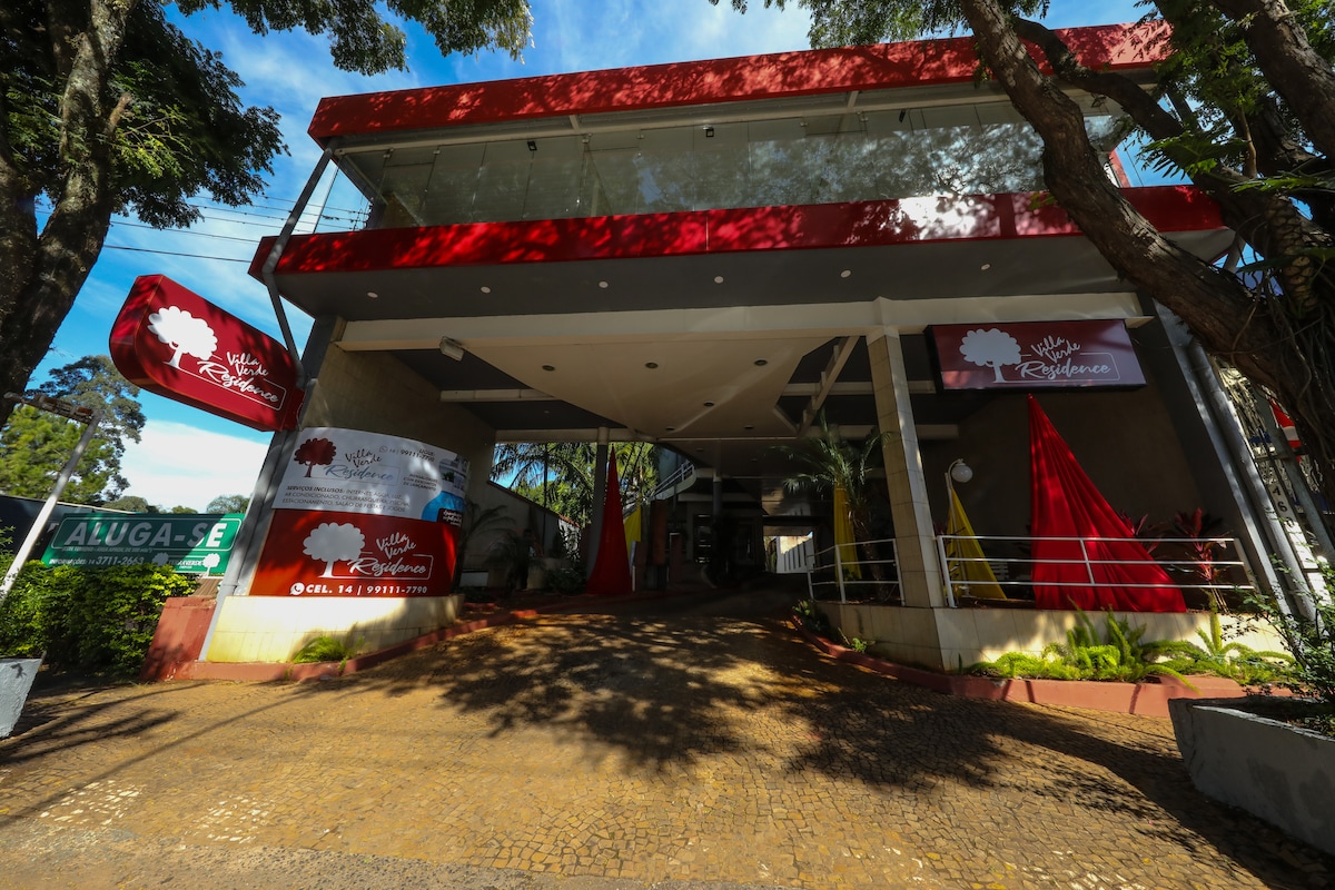 The entrance of Villa Verde Residence is characterized by modern architecture with large glass windows and red accents. A smoothly paved path leads to the entryway, framed by lush greenery and decorative elements. A rental sign is visible on the left side.