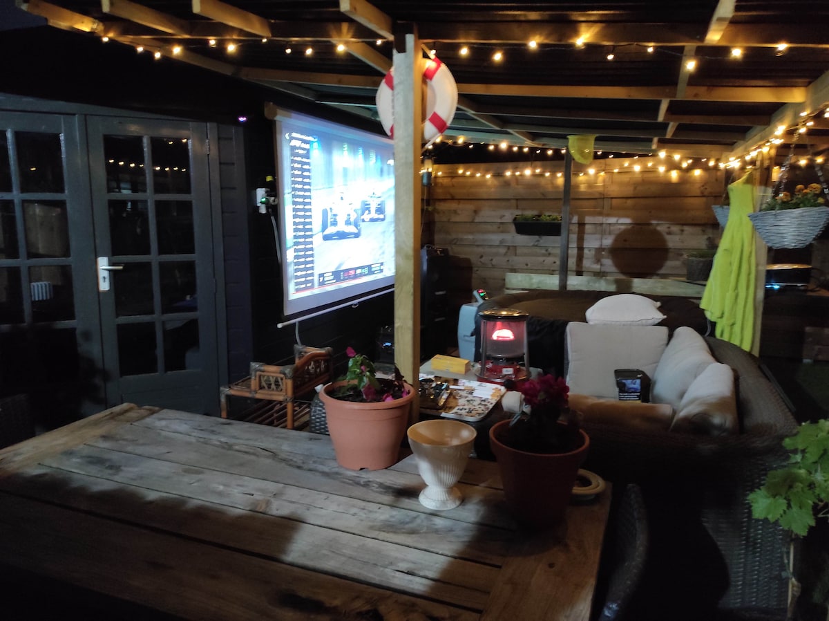 A cozy outdoor space features a wooden dining table adorned with potted plants and a white bowl. A large screen displays entertainment, while string lights create a warm ambiance above. A hot tub can be seen in the background, inviting relaxation.