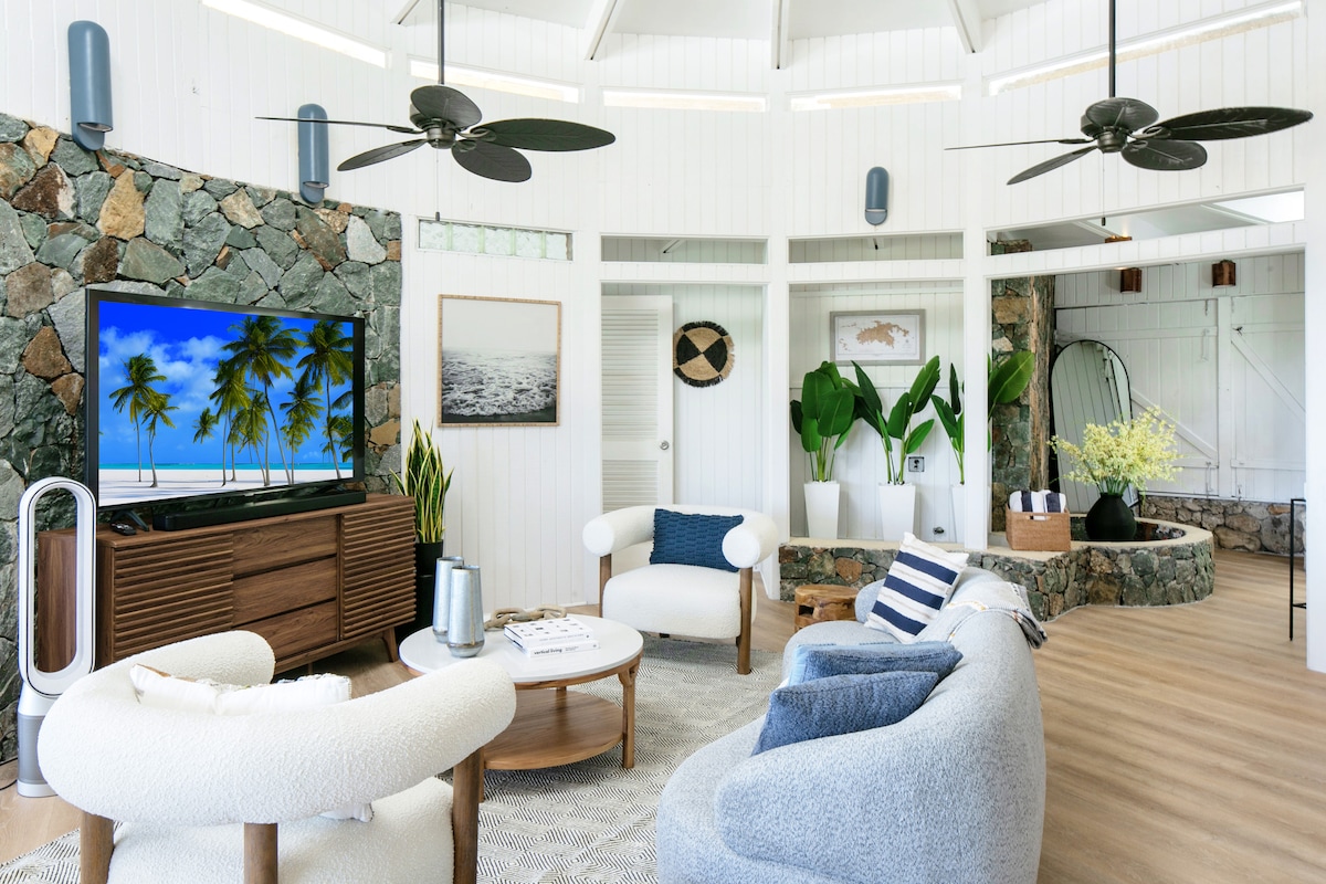 A spacious living area features a large television displaying a tropical scene. The room is accented by a mix of soft seating, including a round chair and a cozy sofa. Natural stone walls provide contrast, while ceiling and portable fans enhance airflow.