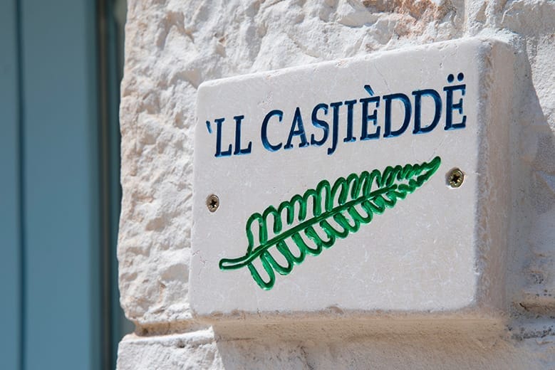 A stone plaque features the name 'Ll Casjèdde' in blue lettering, accompanied by a green fern illustration. The plaque is mounted on a light-colored wall, enhancing the local character of the trullo.