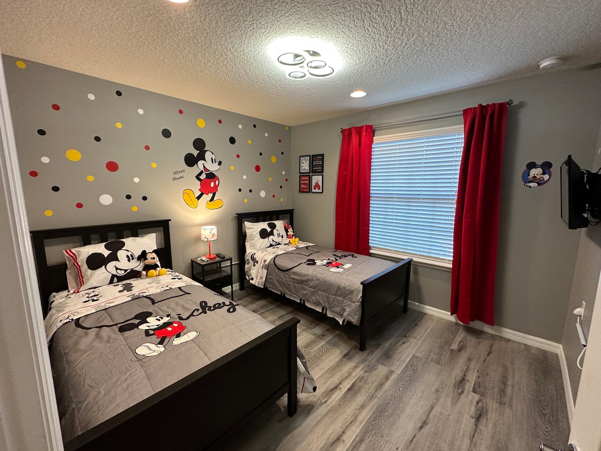 A themed bedroom featuring two twin beds with grey bedding adorned with Mickey Mouse design. The walls are decorated with playful polka dots and Mickey illustrations. Red curtains frame the window, and a bedside lamp adds a cozy touch, completing the fun atmosphere.