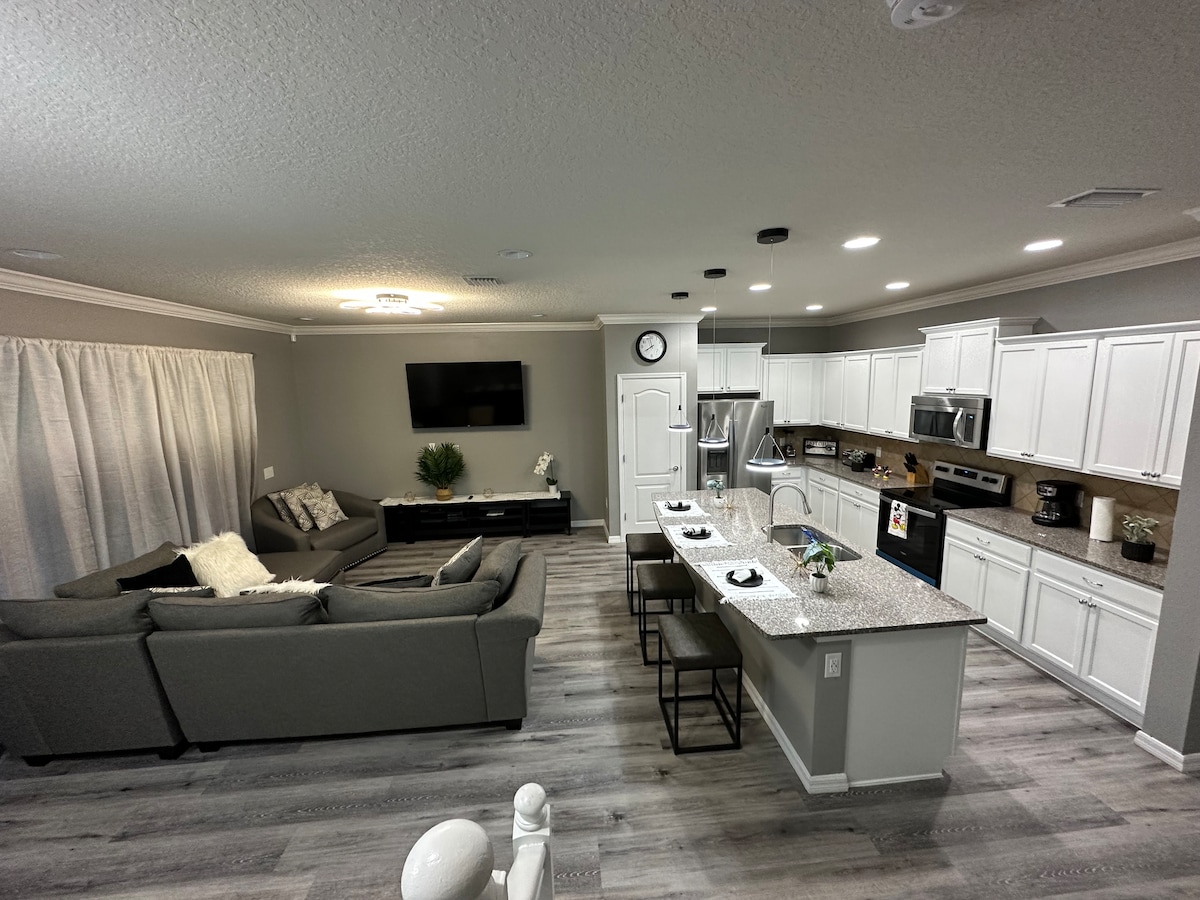 An open-concept living area is highlighted by a spacious gray sofa facing a flat-screen TV. The kitchen features a large island with seating for three, equipped with modern appliances, and light-colored cabinetry. Natural light flows through the window drapes, creating an inviting space.