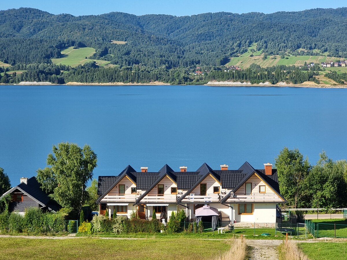 A row of modern houses with black roofs is set against a backdrop of rolling green hills and calm blue waters. The serene lake reflects the sky, while lush trees frame the scene, creating a peaceful atmosphere ideal for relaxation.