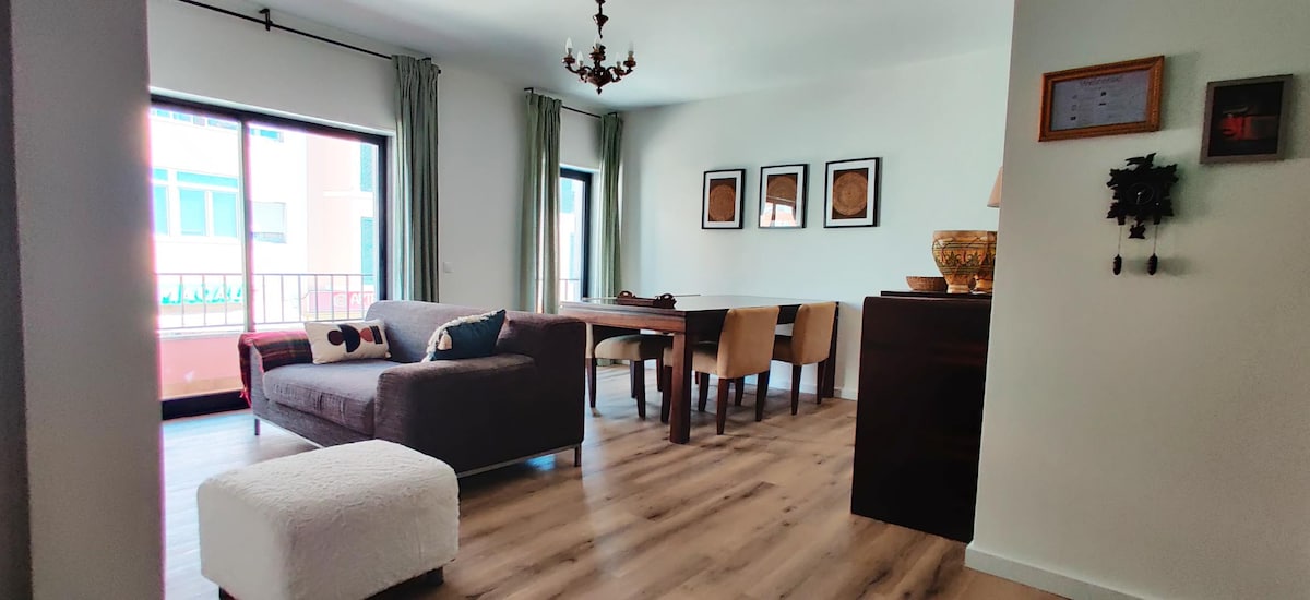 A spacious living area is visible, featuring a comfortable sofa and a wooden dining table surrounded by chairs. Large windows allow natural light to fill the room, complemented by light curtains. Decorative items and artwork are displayed on the walls, adding a personal touch.