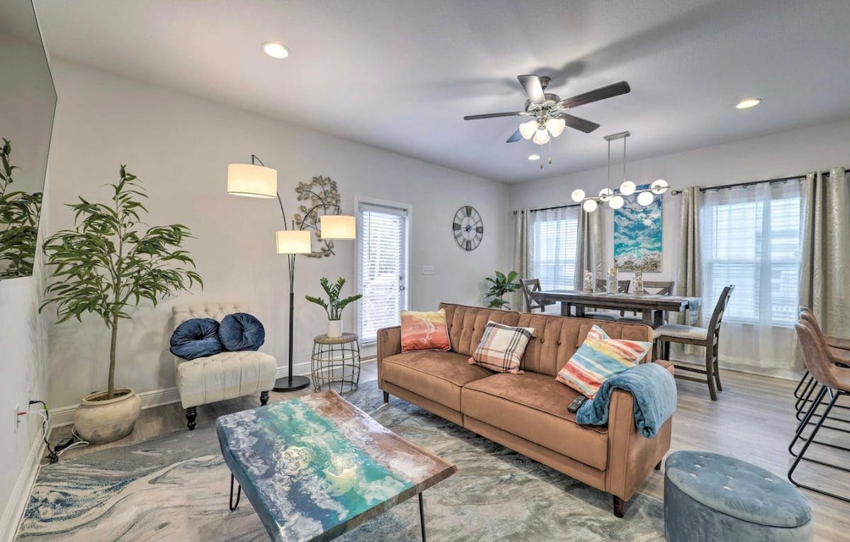 A welcoming living area features a brown sofa adorned with colorful pillows, set against a light-toned wall. A decorative coffee table rests on a large area rug. A ceiling fan and various light sources illuminate the space, complemented by a large dining table visible in the background.