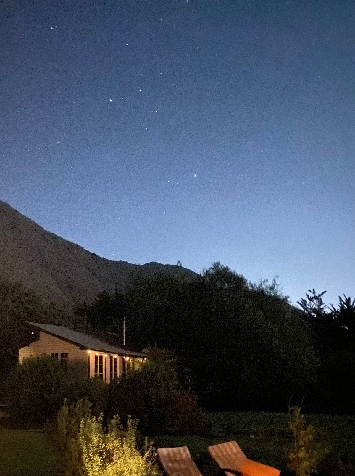 The image depicts a serene night scene with a cottage nestled against a mountainous backdrop. Stars are visible in the clear sky above, while the faint glow of the cottage's exterior lights softly illuminates the surrounding landscape. A pair of lounge chairs can be seen in the foreground.