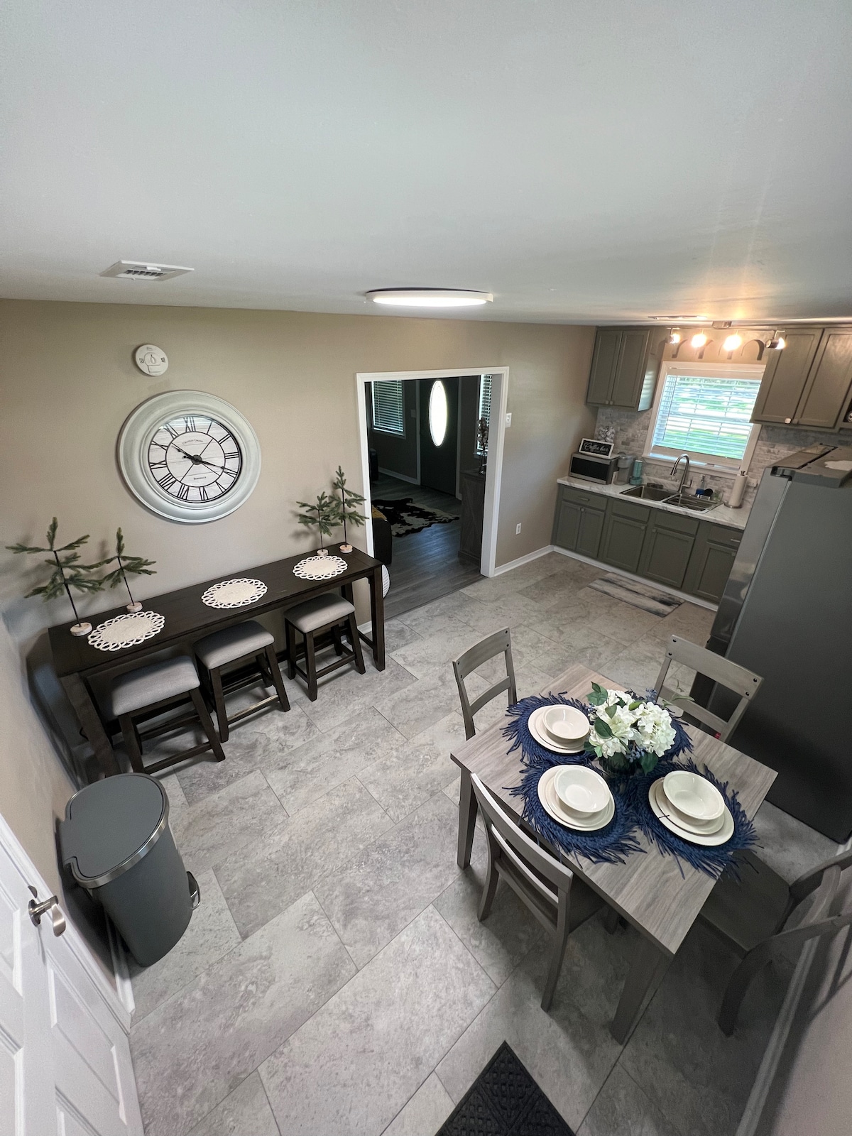 An inviting dining area is depicted, featuring a table set for four with white plates and a blue centerpiece. A large wall clock hangs above the table, and soft lighting illuminates the space. Gray cabinetry and modern appliances are visible in the background.