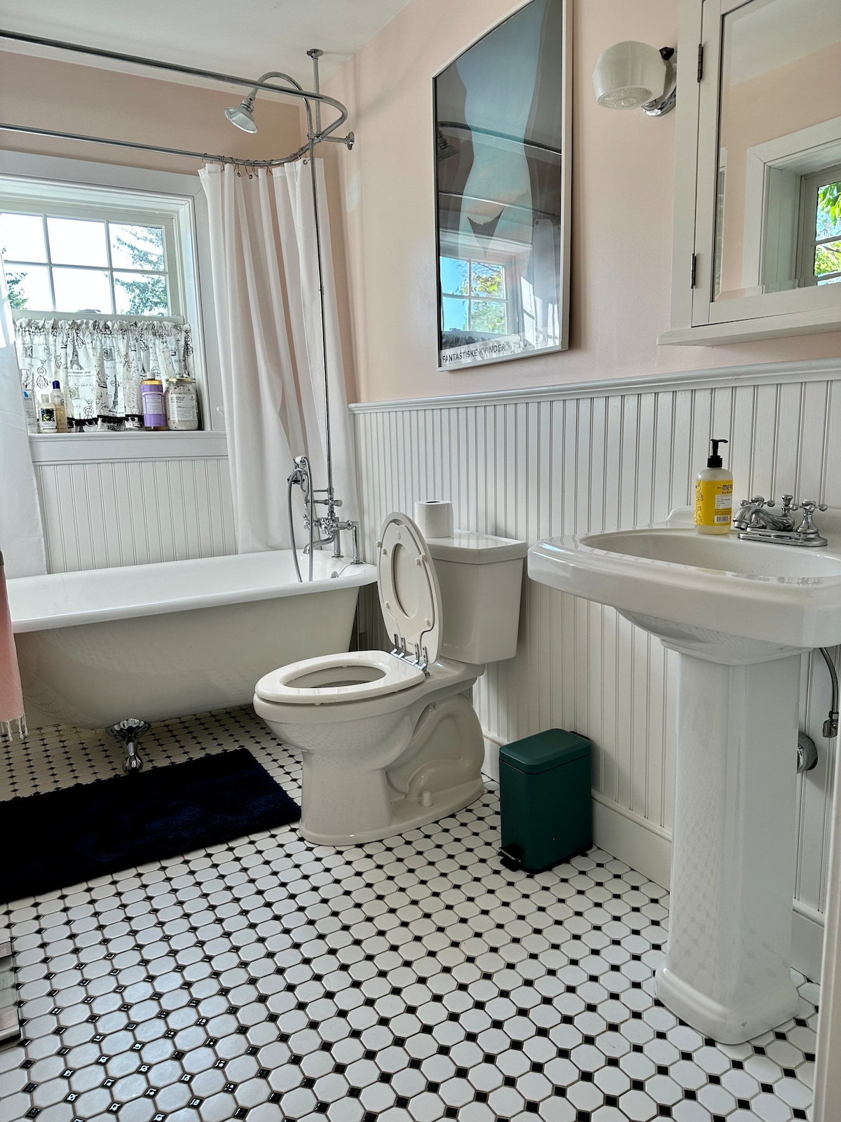 A full bathroom features a clawfoot tub, a pedestal sink, and a toilet. White tiles with black accents cover the floor. A large mirror hangs above the sink, and light enters through a nearby window dressed with a sheer curtain.