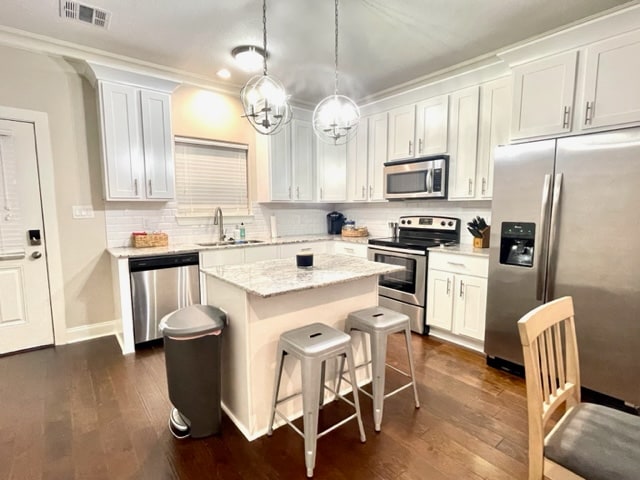 A modern kitchen features white cabinetry and a central island with granite countertops. Stainless steel appliances, including a refrigerator and oven, are present. Three metal stools are positioned at the island, and a trash can is discreetly located nearby.
