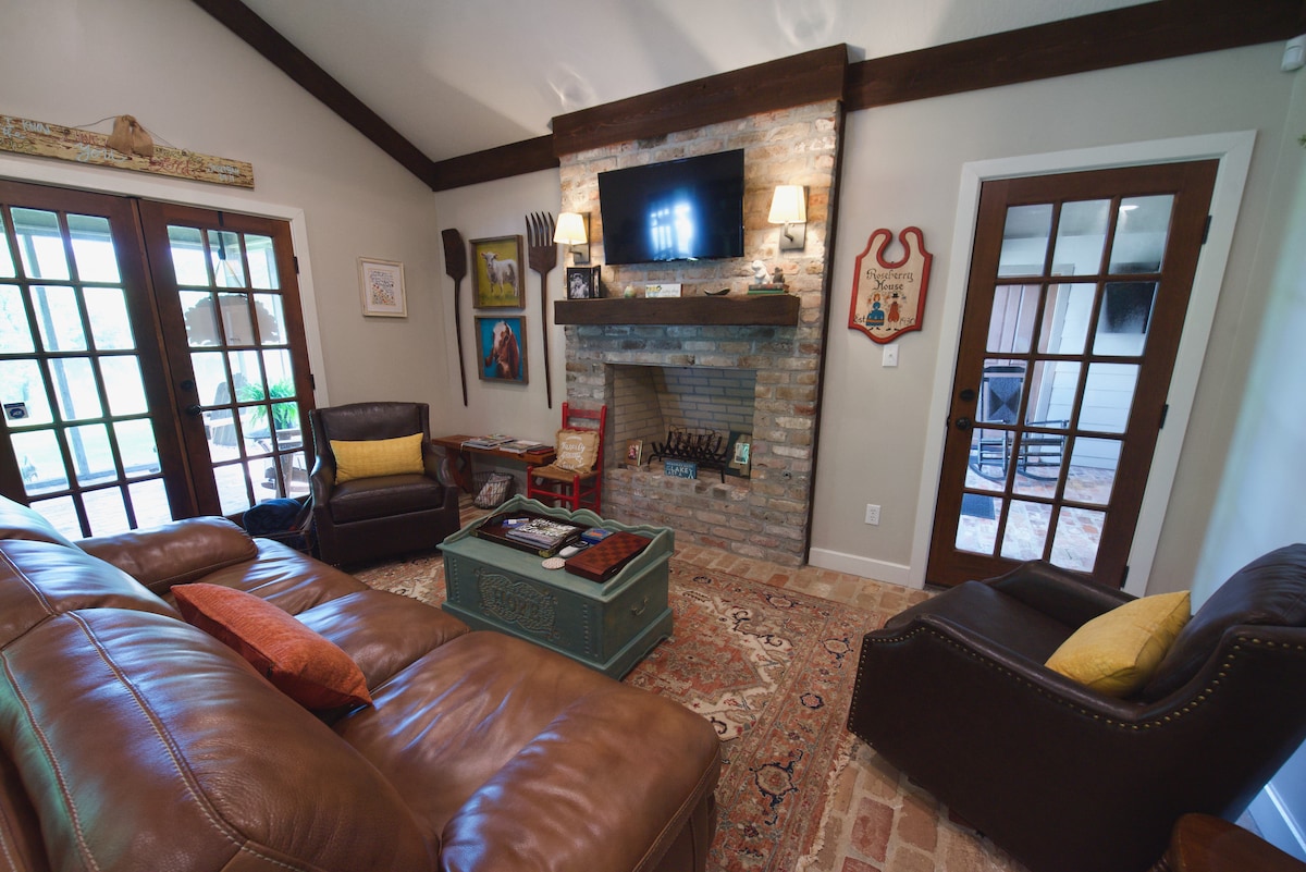 A warm living area features a comfortable leather sofa and chairs, arranged around a rustic coffee table. A brick fireplace with a TV above serves as a focal point. French doors lead to an outdoor space, while natural light brightens the room.
